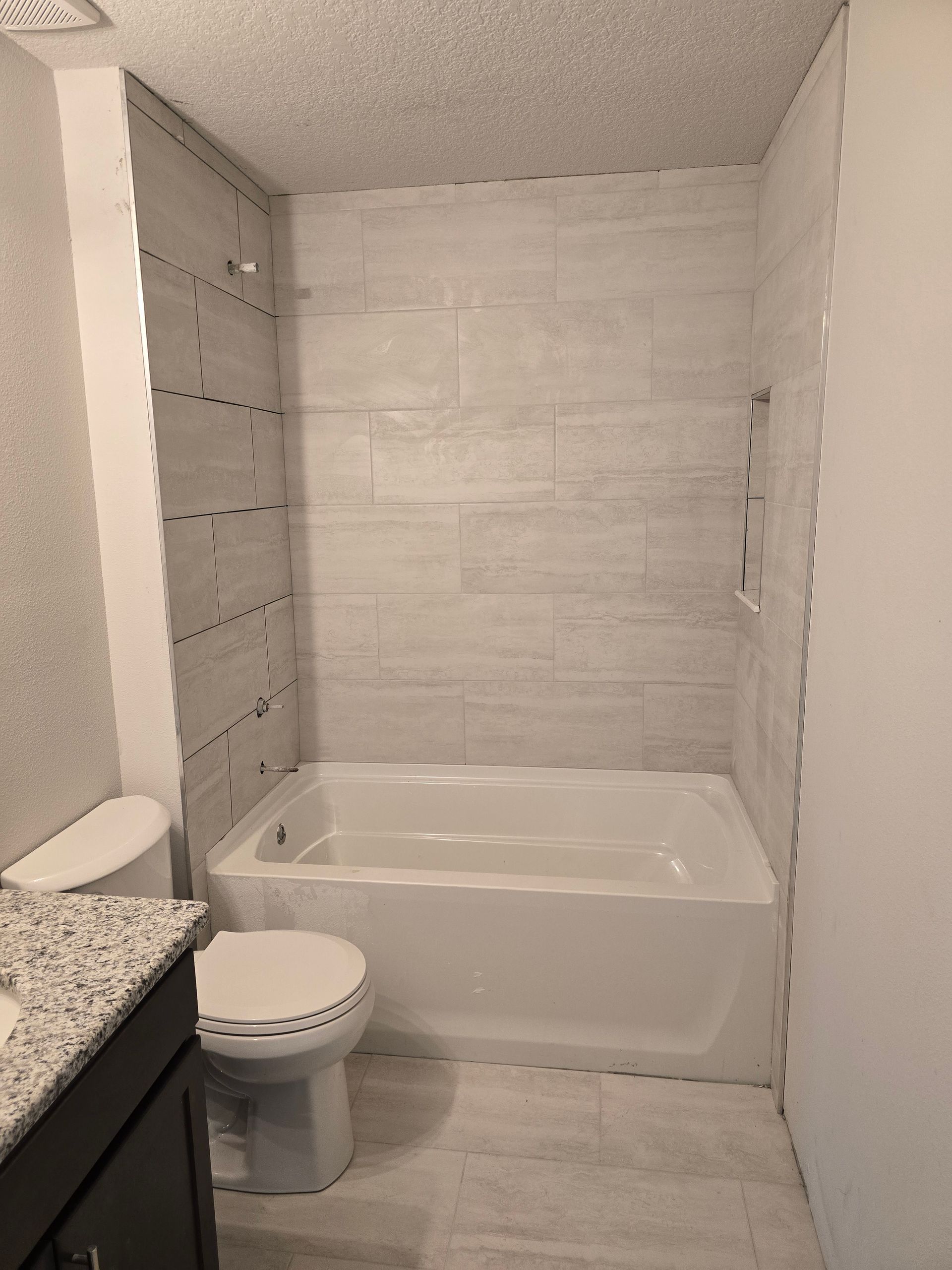 Small bathroom with a white bathtub, toilet, dark vanity, and light tile shower wall.