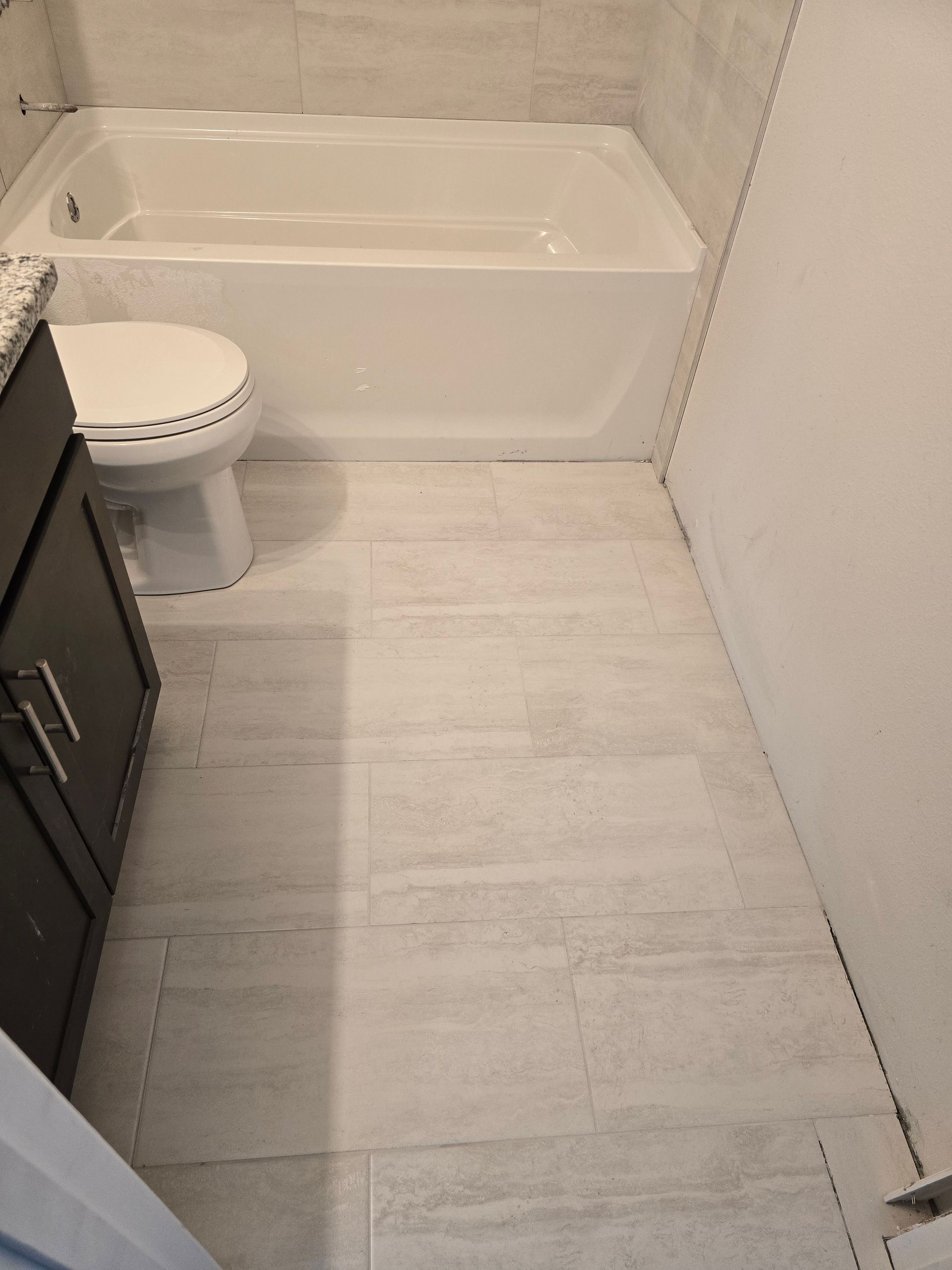 Narrow bathroom with tub, toilet, dark vanity, and light wood-look tile floor.