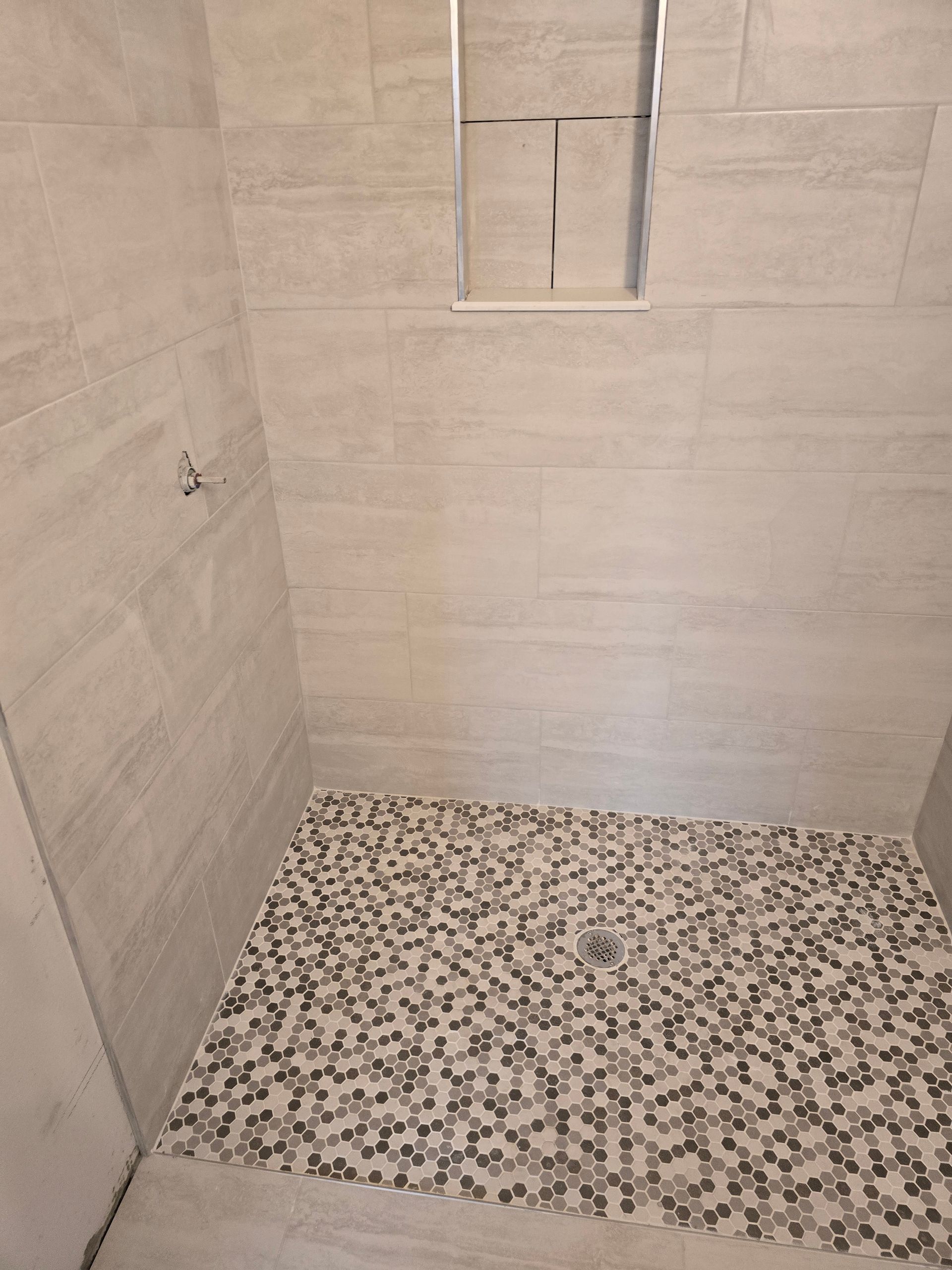 Empty tiled shower with beige walls and pebble-pattern floor, showing a small window.