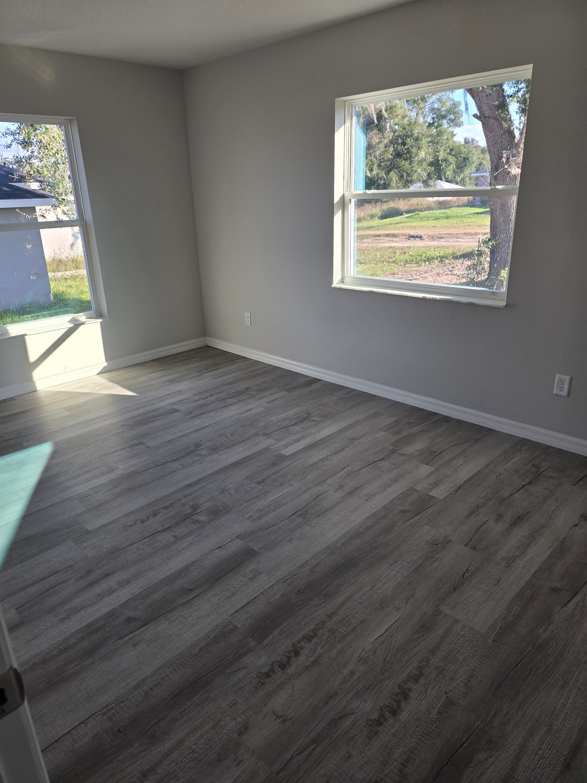 Empty room with gray wood-look flooring, white walls, and two windows letting in daylight.
