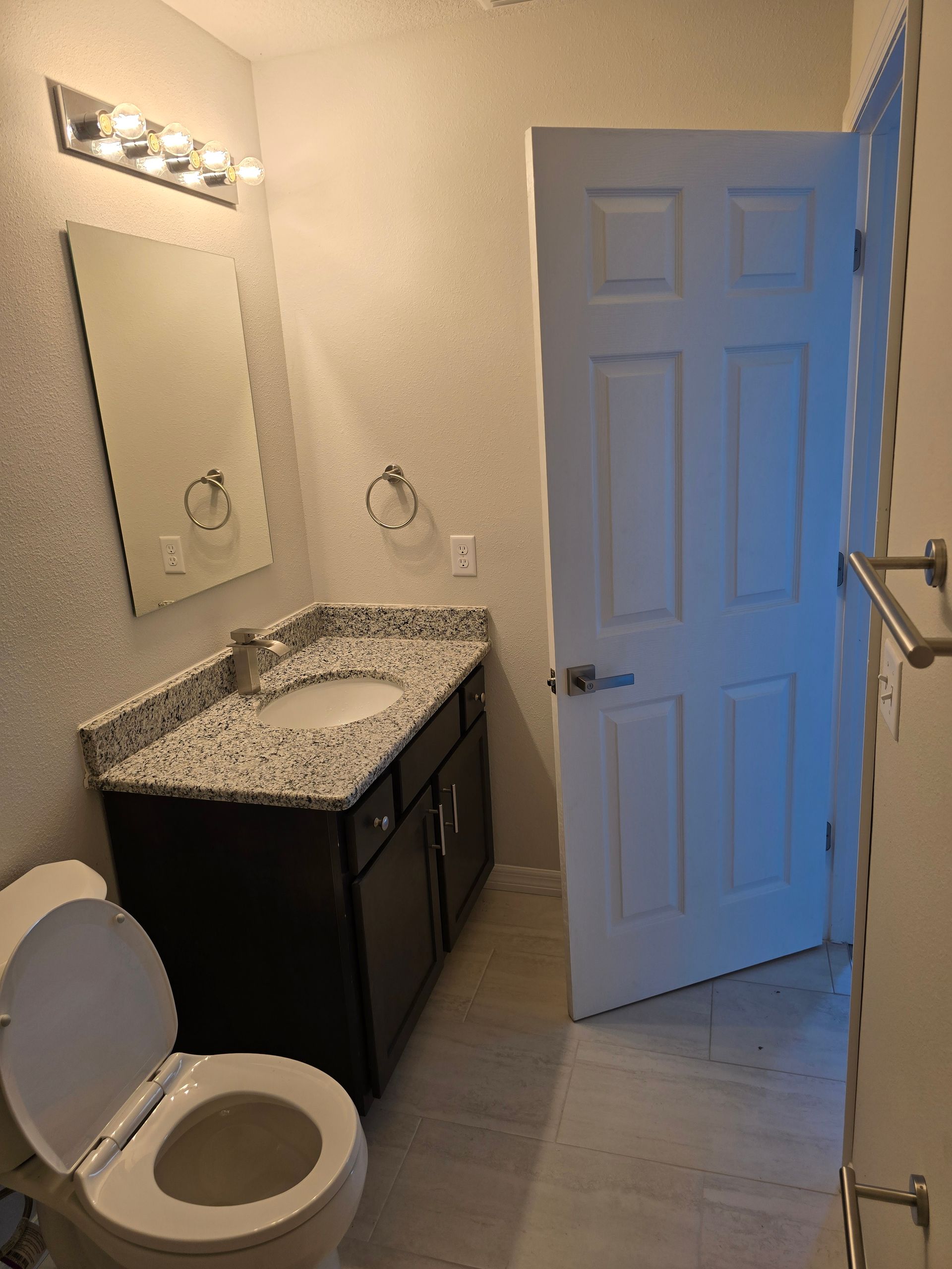 Small bathroom with toilet, granite vanity, mirror, and open door leading to a hall.