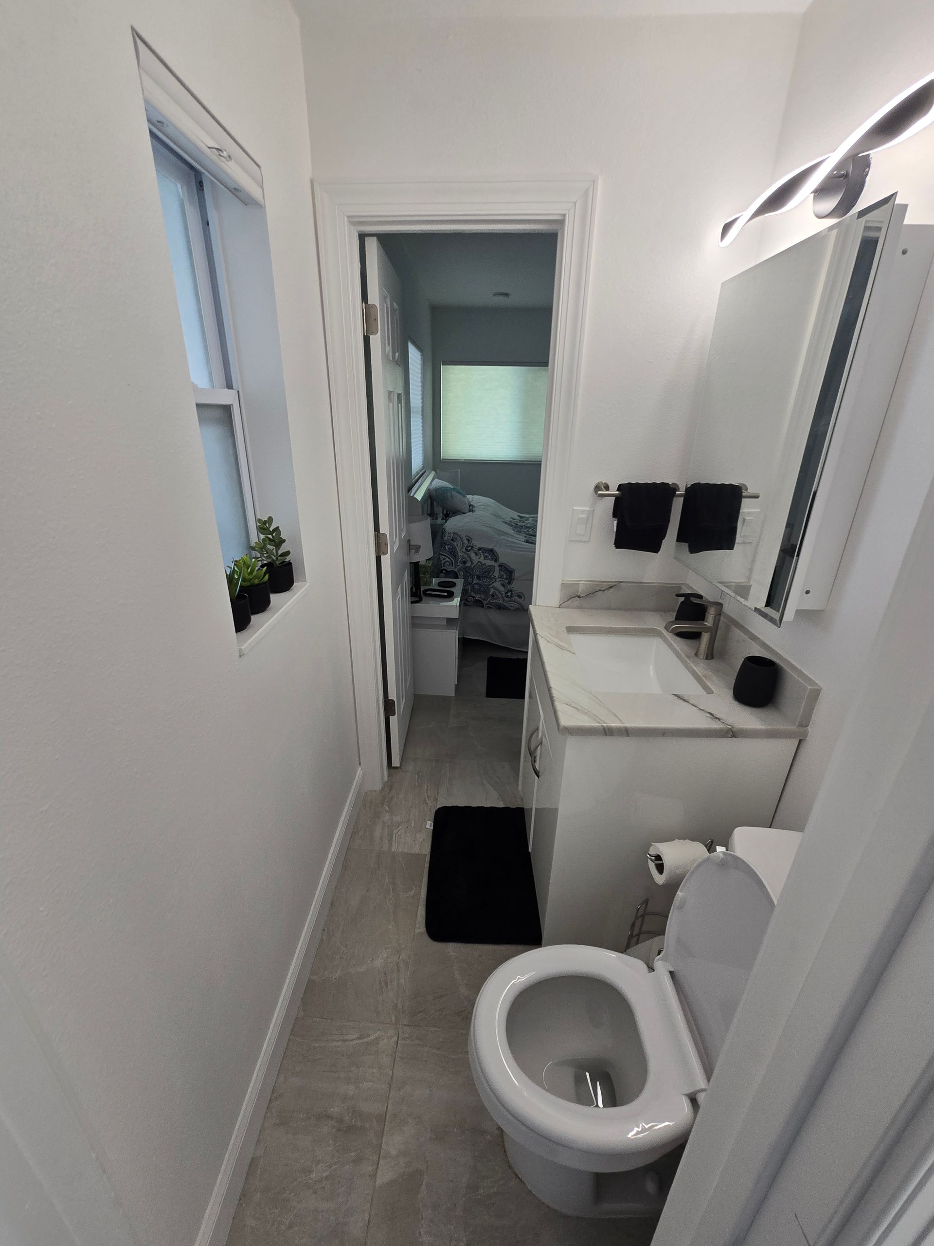 Narrow white bathroom with sink, toilet, mirror cabinet, and doorway to a bedroom beyond.