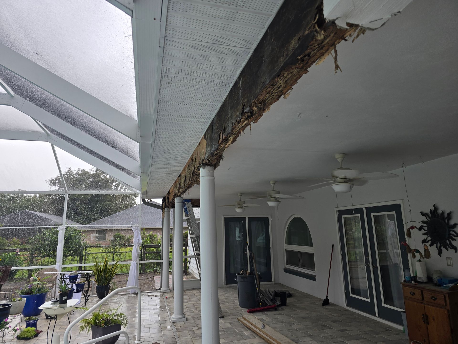 Damaged porch roof with exposed wood and rust, beside a white house with columns and potted plants.