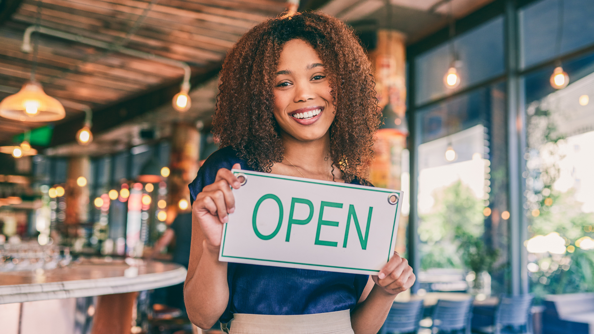 Person holding an “OPEN” sign inside a warmly lit café.