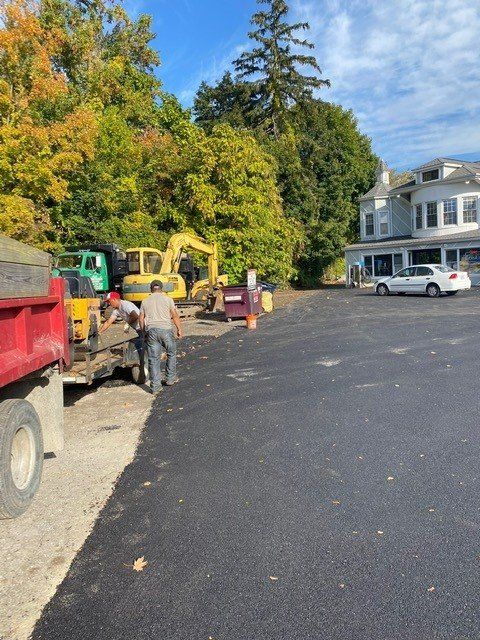 Construction equipment and workers on a paved driveway beside a white house and autumn trees.