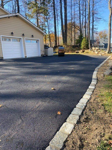 Fresh black asphalt driveway beside a garage, bordered by a light stone curb and trees in autumn.