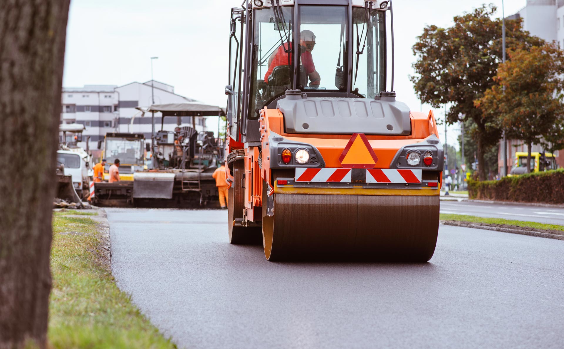 Orange road roller paving a wet street, with other construction vehicles behind it.