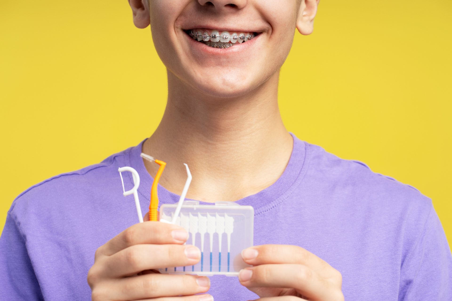 Person with braces holding dental cleaning tools, smiling against a yellow background. Person with braces holding dental cleaning tools, smiling against a yellow background.