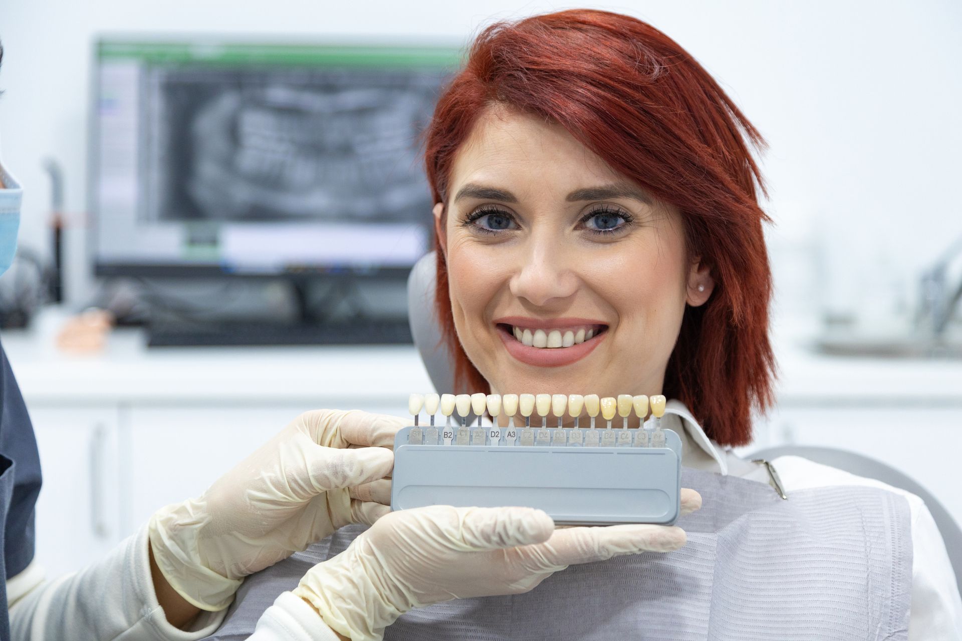 Woman in dental chair smiles while dentist holds teeth shade guide. Clinic setting.