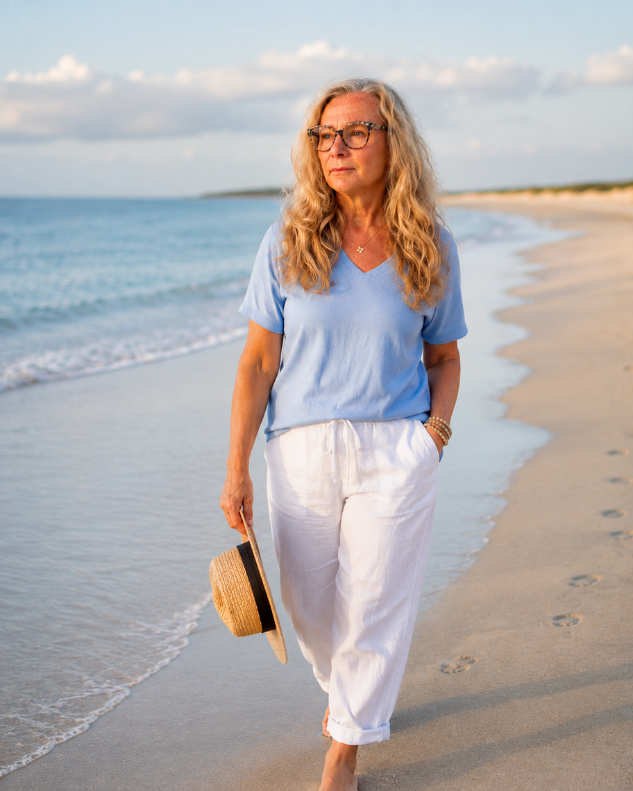 Woman walking barefoot along a sunny beach beside calm blue water, wearing a navy shirt and white shorts