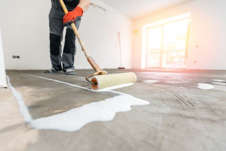 Person rolling white floor coating onto a concrete floor in a bright room
