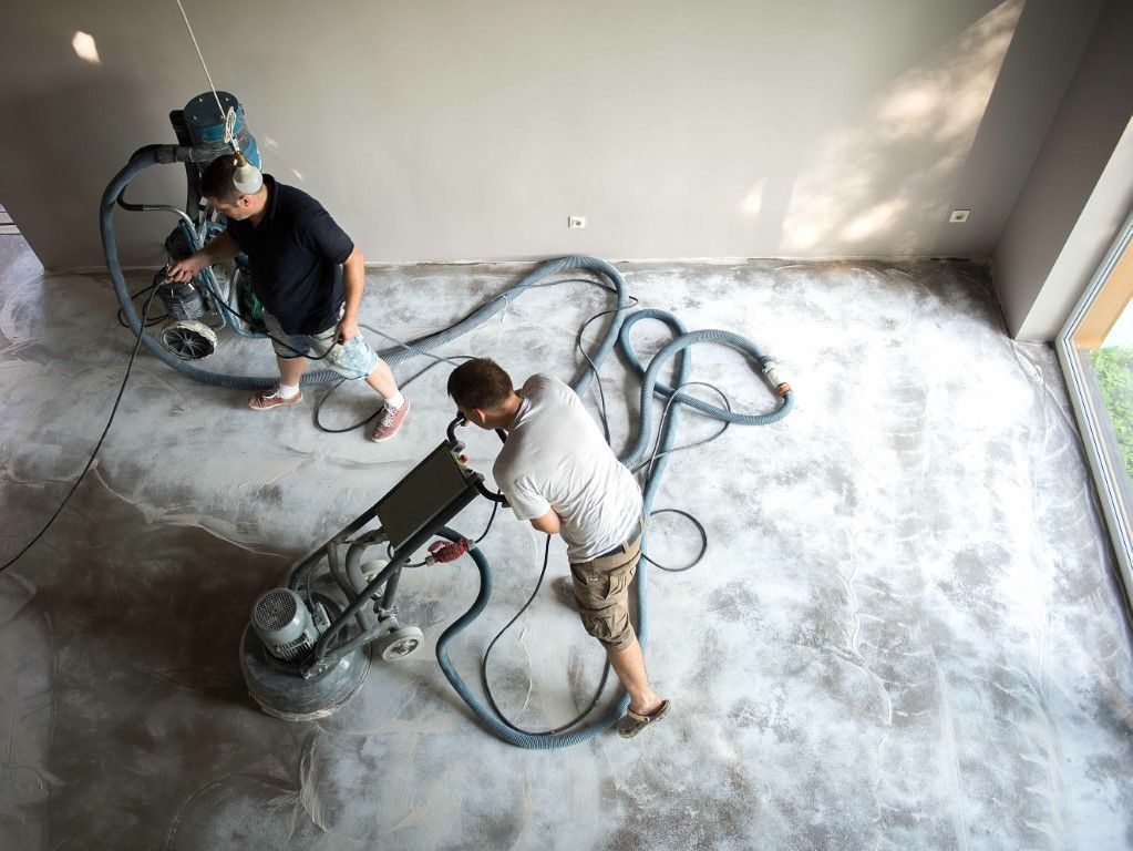 Two workers sanding a concrete floor with floor grinders in an empty room