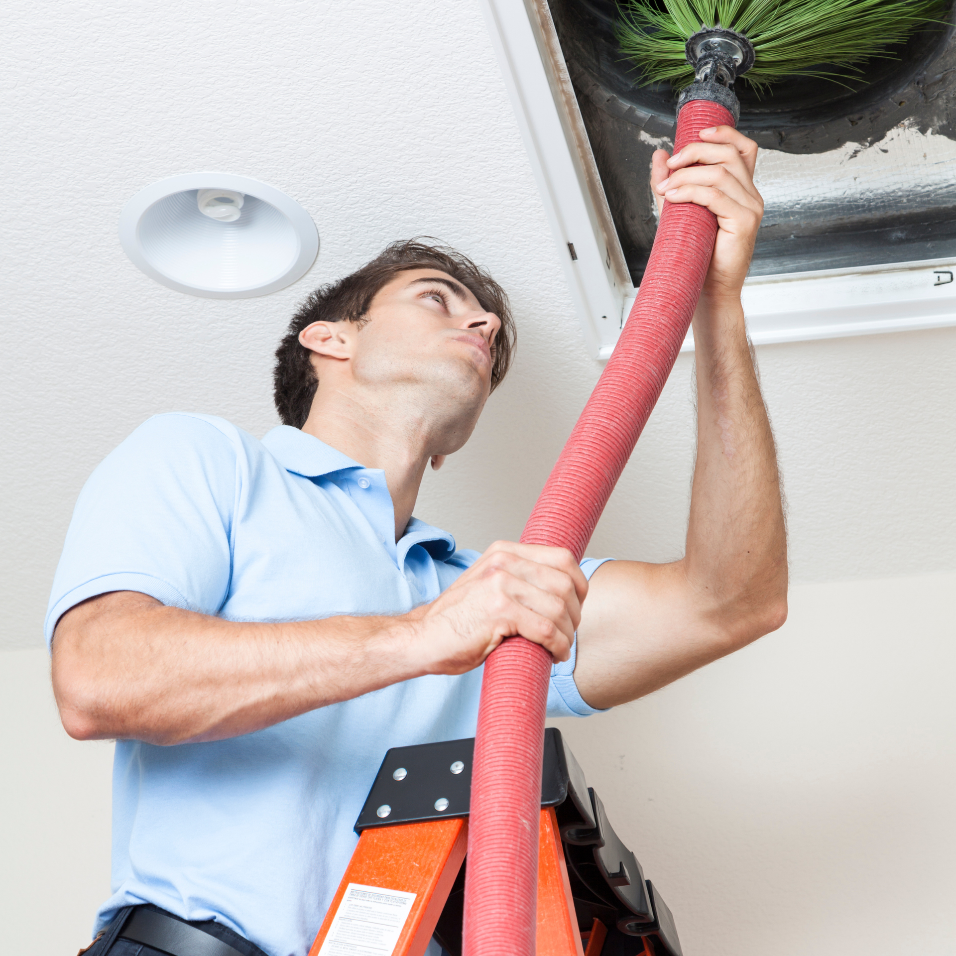 Man cleaning air duct with hose, standing on a ladder in a white ceiling.