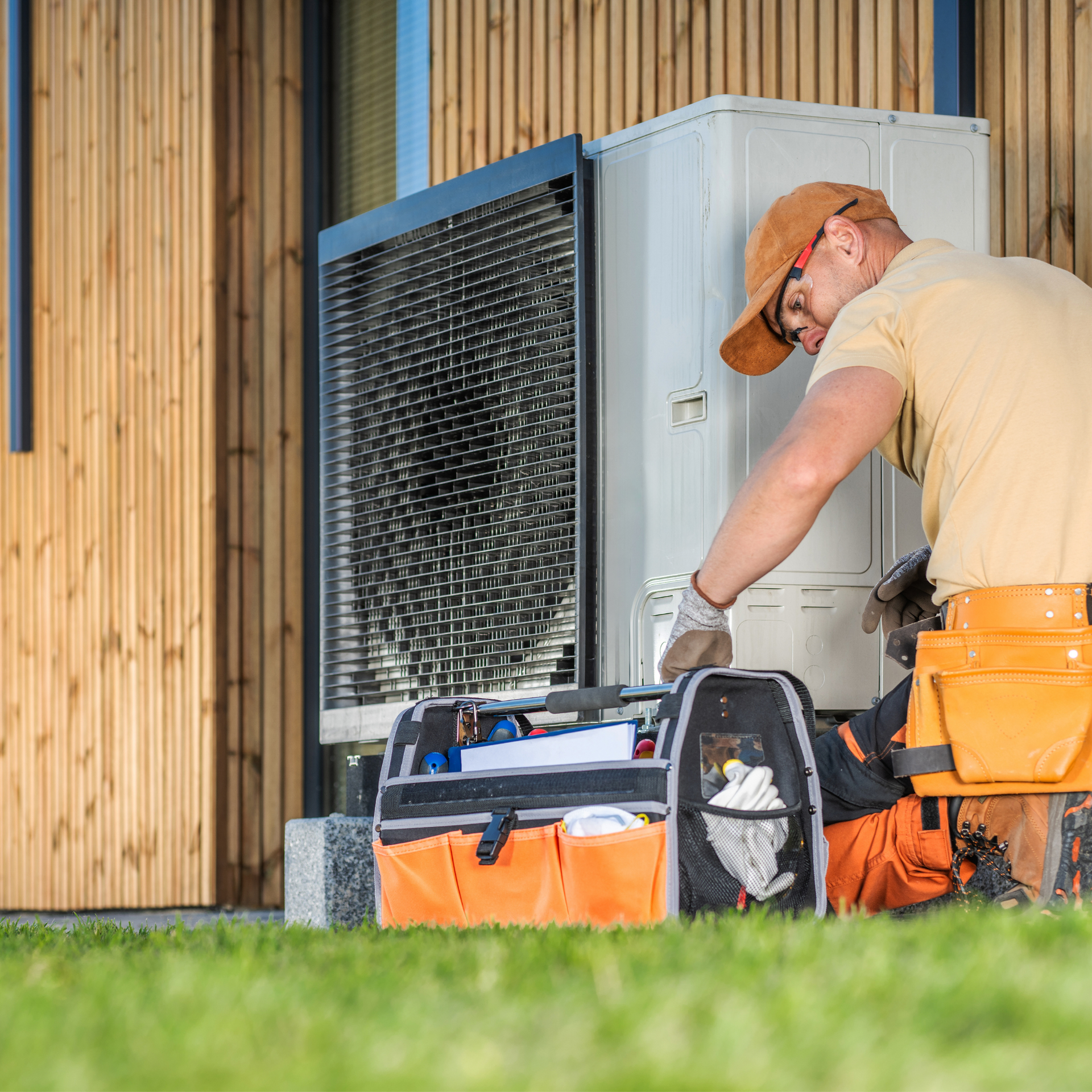 Technician working on an outdoor HVAC unit; tool bag on the ground, wooden building in the background.