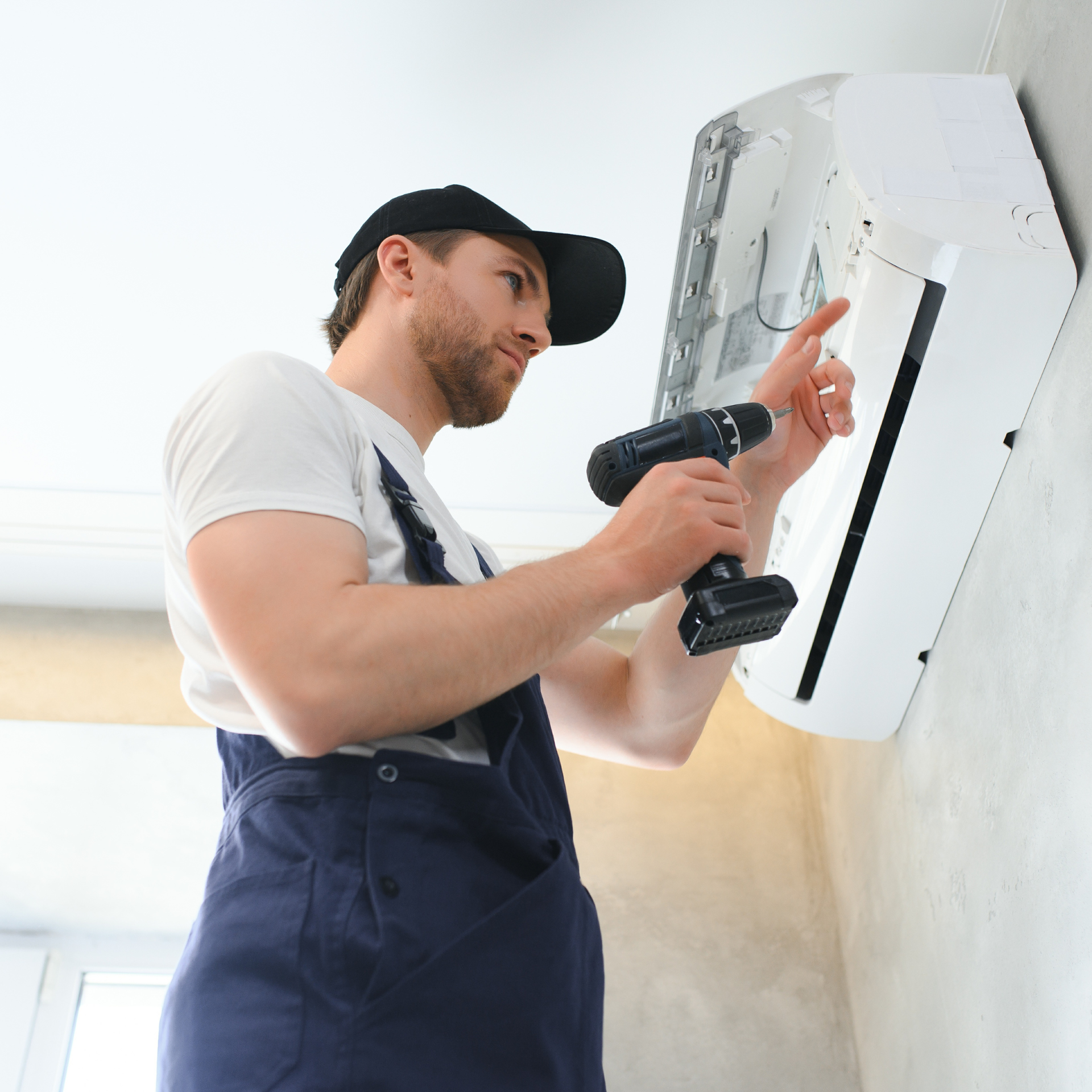 Man in overalls installs an AC unit with a drill. White unit on wall, indoor setting.