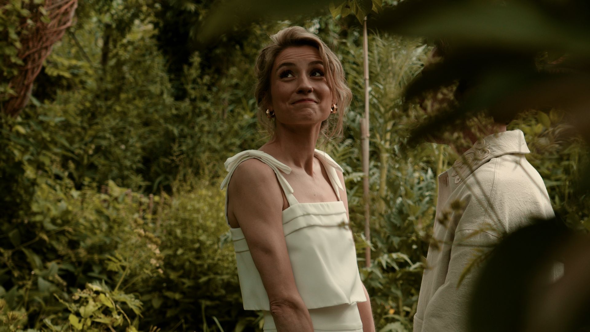 Woman in a white dress smiling in a lush garden, with a white statue in the background
