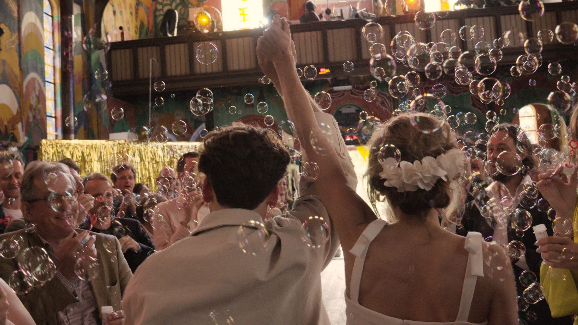 Newlyweds raise their arms amid falling bubbles at a lively indoor celebration