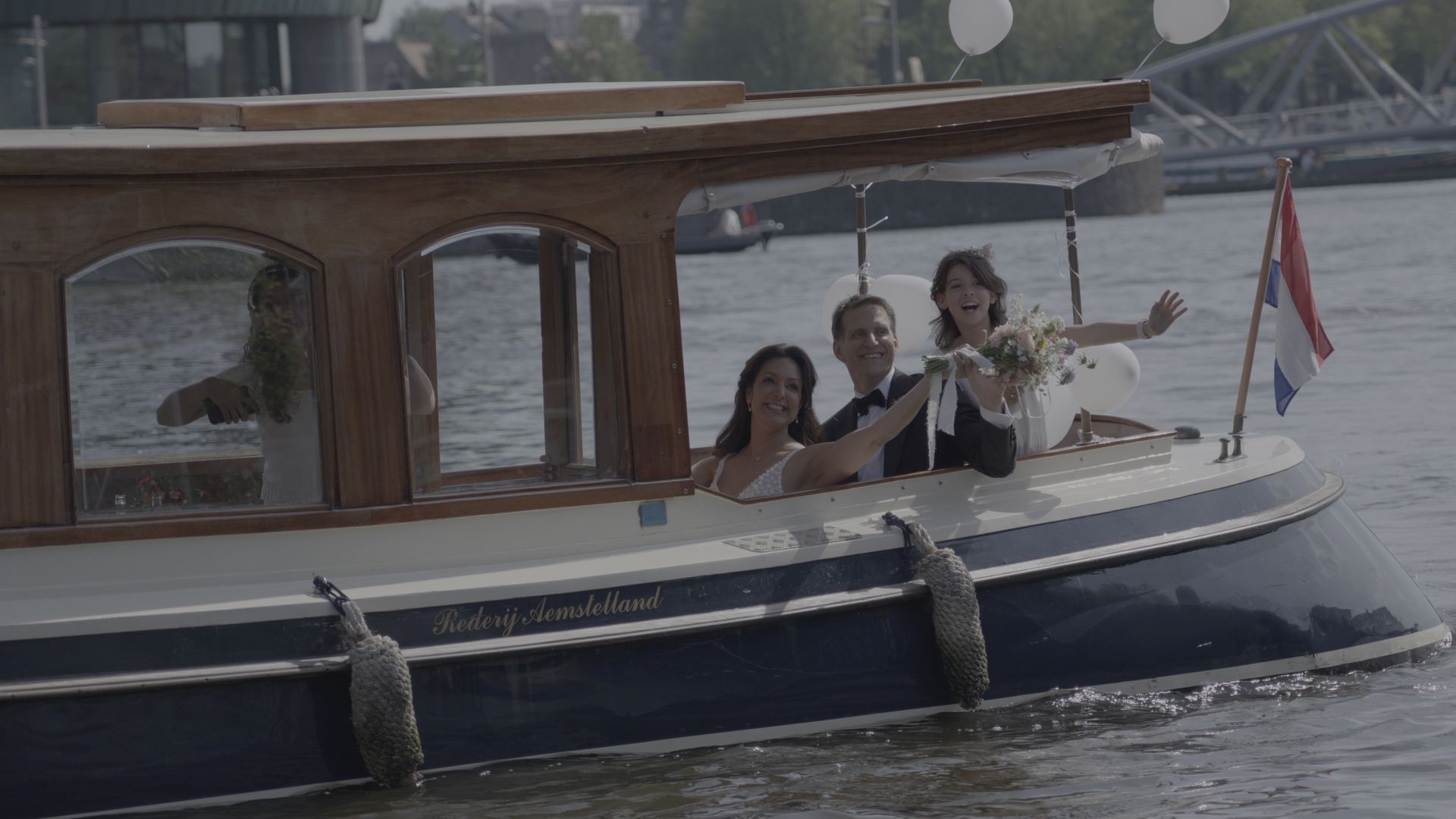 Two people waving from a small wooden boat on calm water, with a red flag at the stern.
