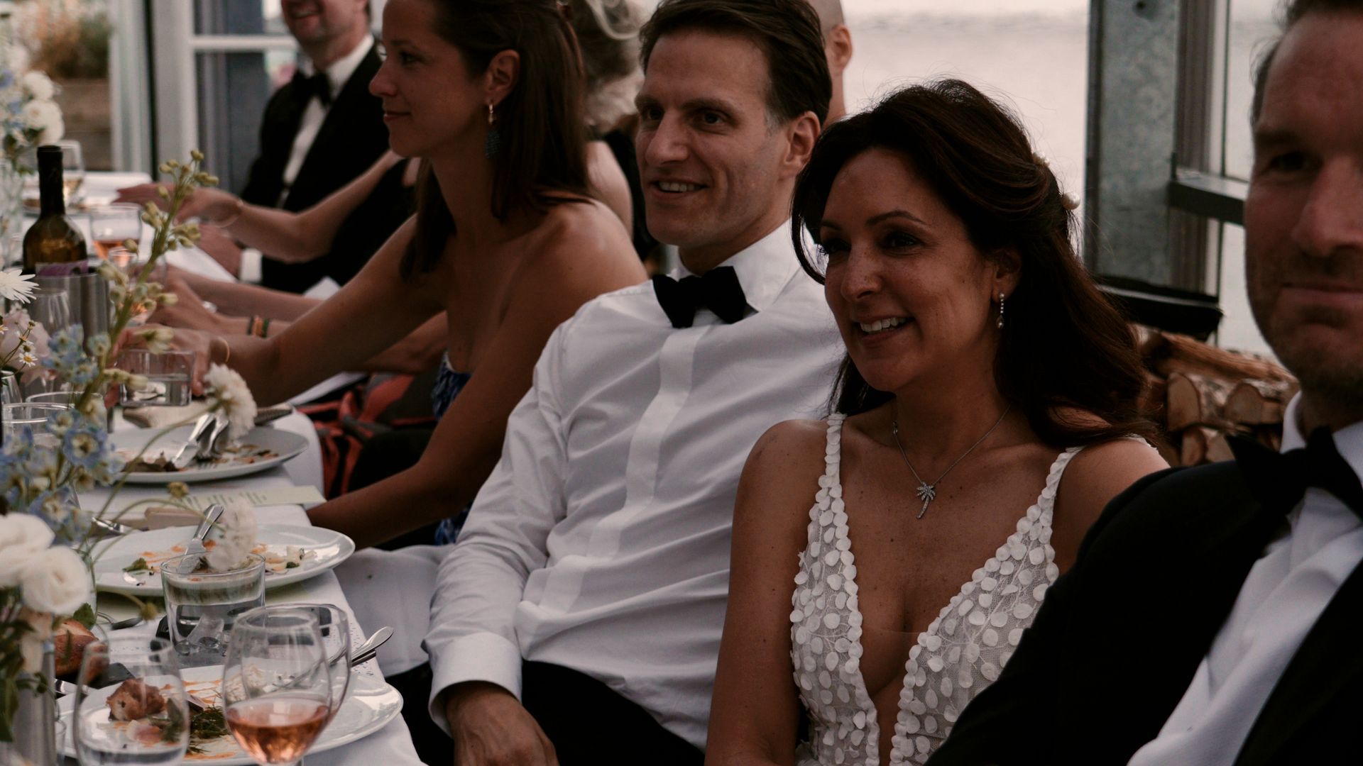 Guests in formal attire seated at a long banquet table outdoors, smiling and dining together