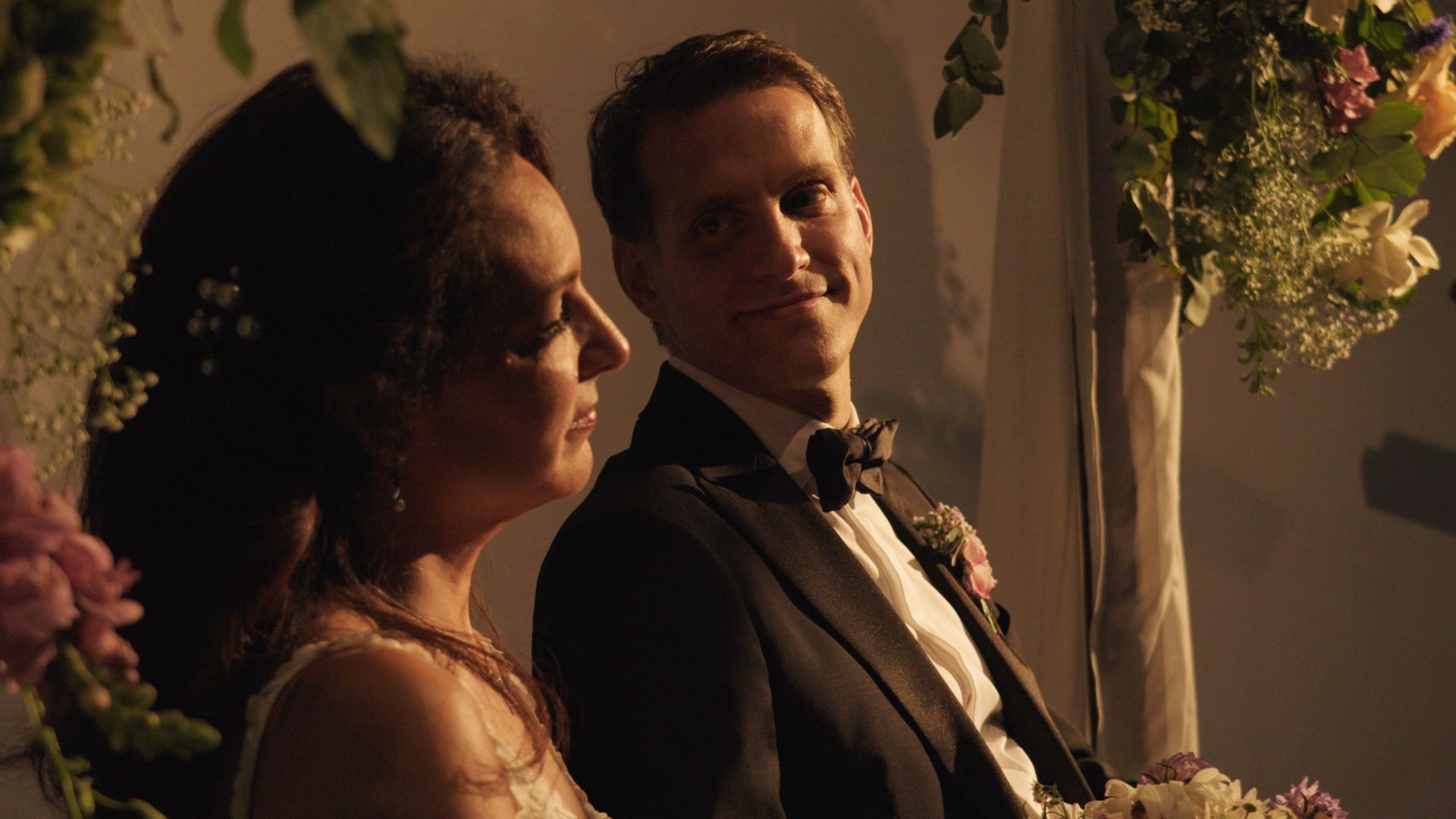 Two people in formal wear smiling at a softly lit floral wedding reception