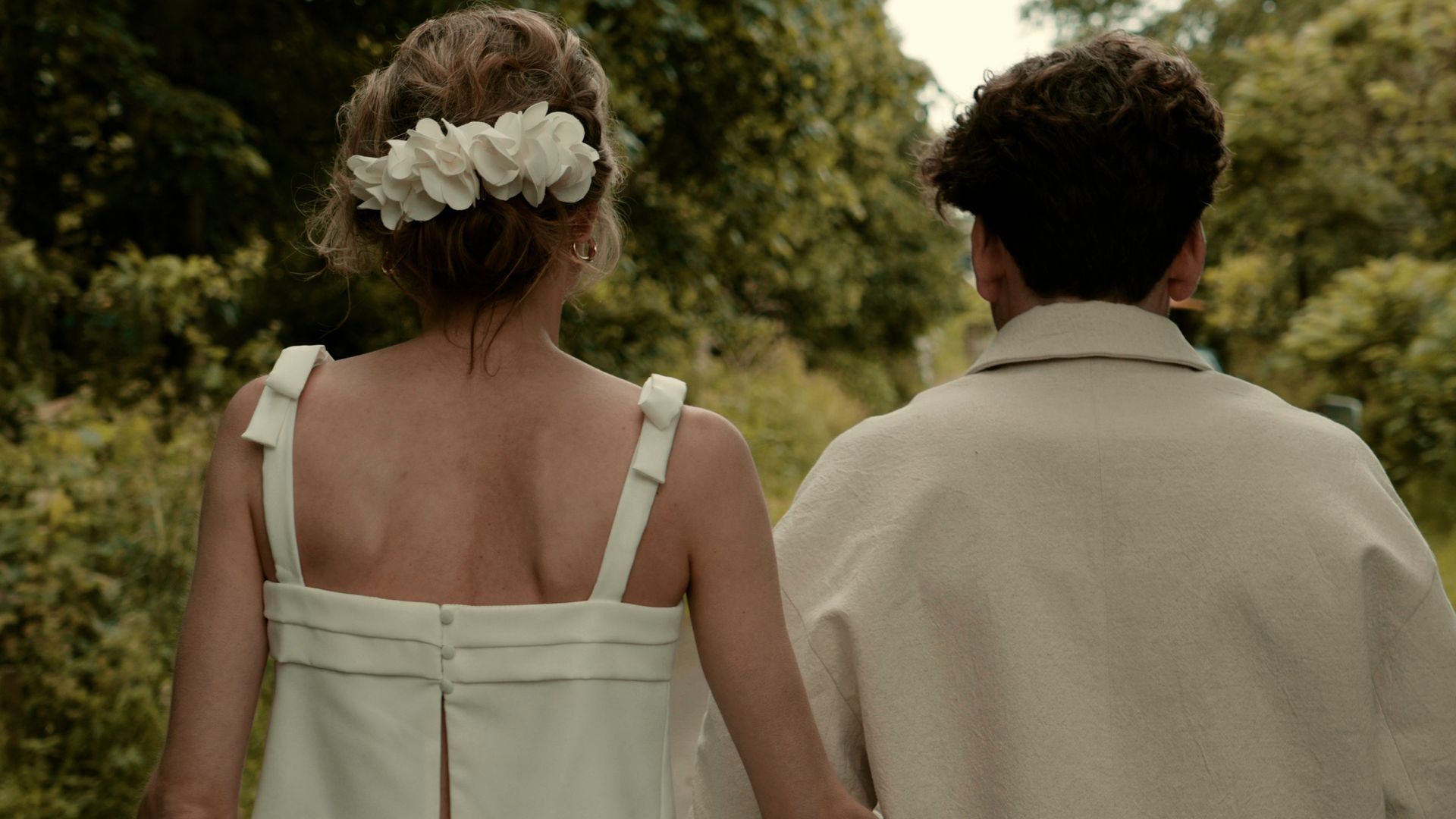 Two people in light clothing walking away through a garden, one with flowers in their hair.