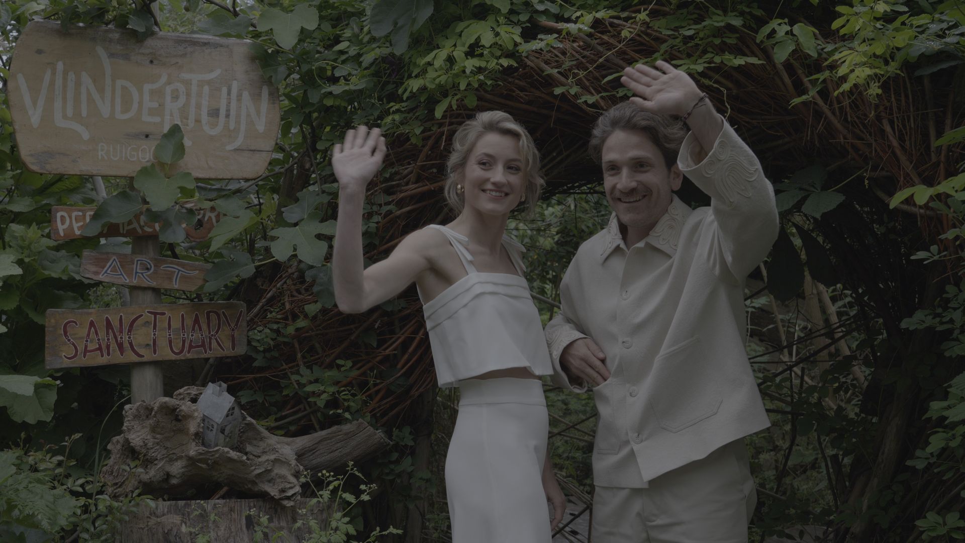 Two people waving in front of a vine-covered sign in a lush garden setting.