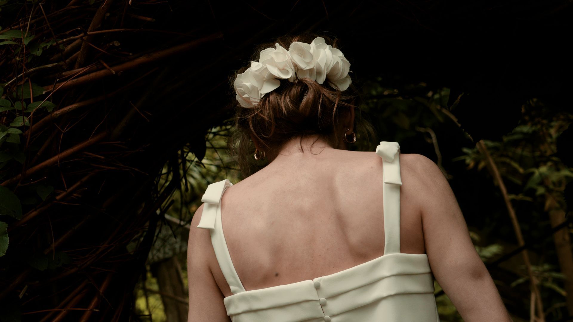 Back view of a person in a white dress and flower crown in a dark garden path