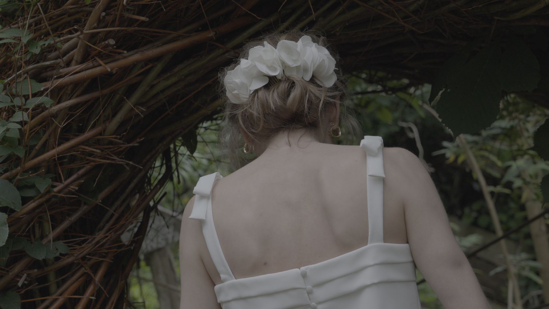 Woman in a white dress seen from behind, walking beneath a leafy arch outdoors.