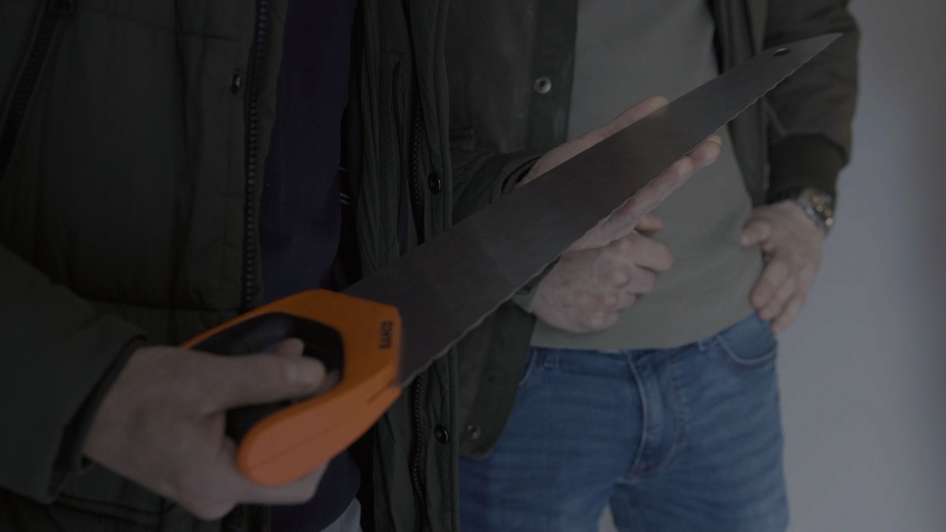 Close-up of hands holding a black-and-orange handsaw beside a person in a jacket and jeans