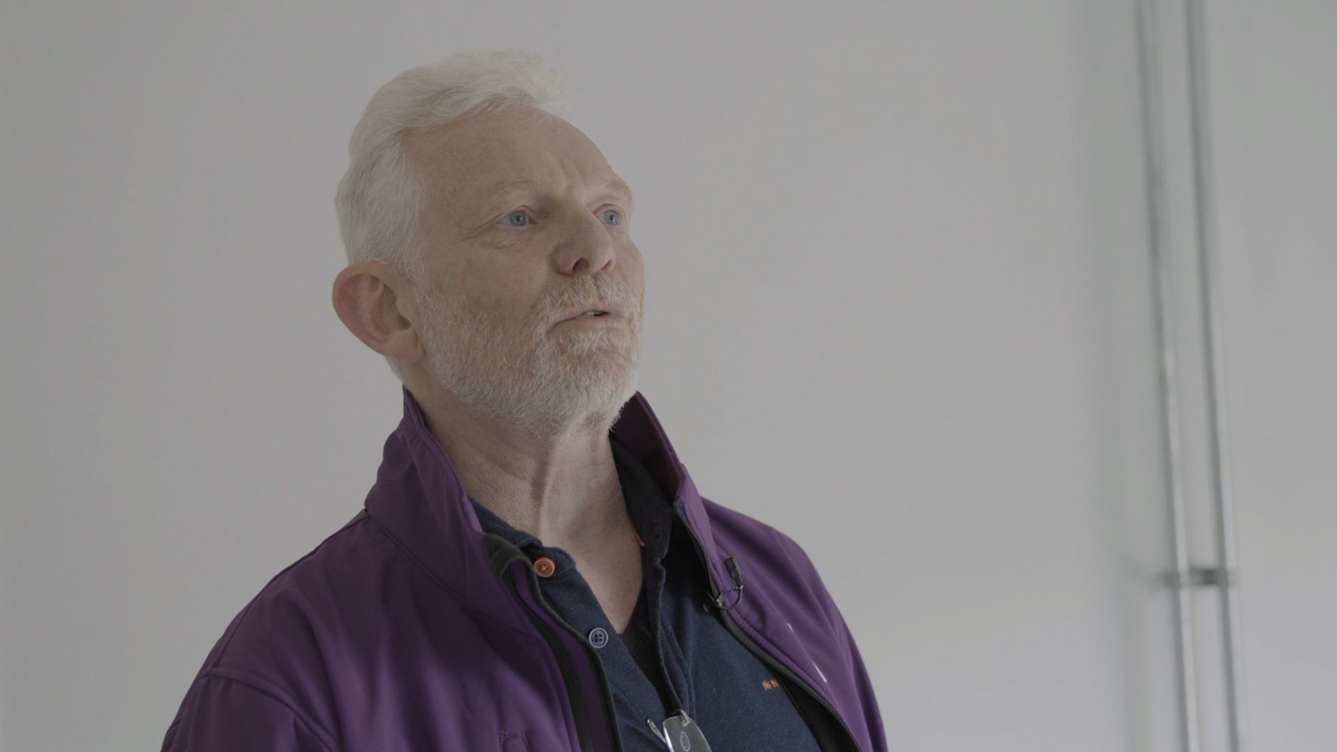 Older man in a purple jacket standing against a plain white wall, looking upward thoughtfully
