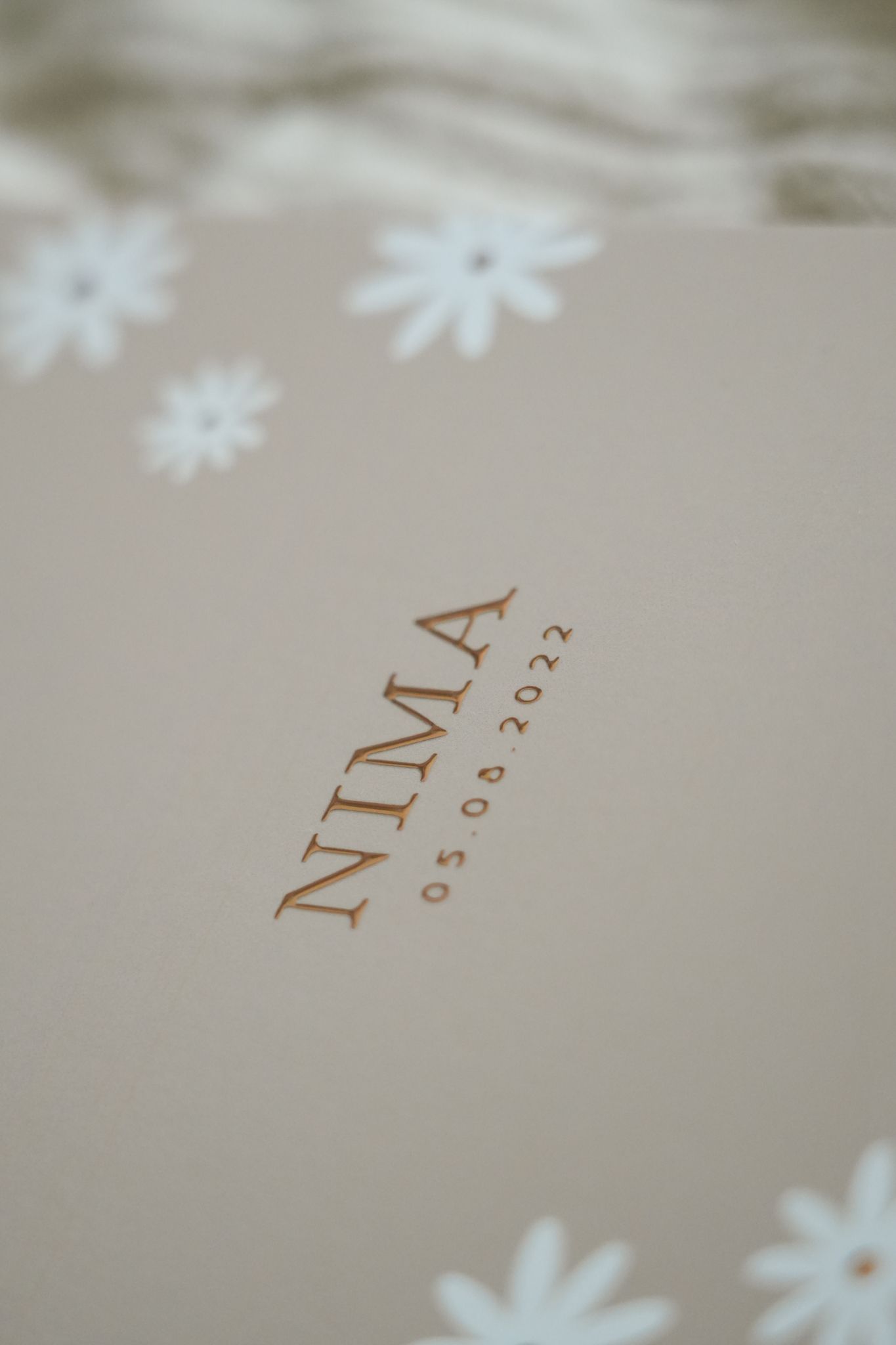 Close-up of a cream card with brown “NIMA” text and white floral decoration.