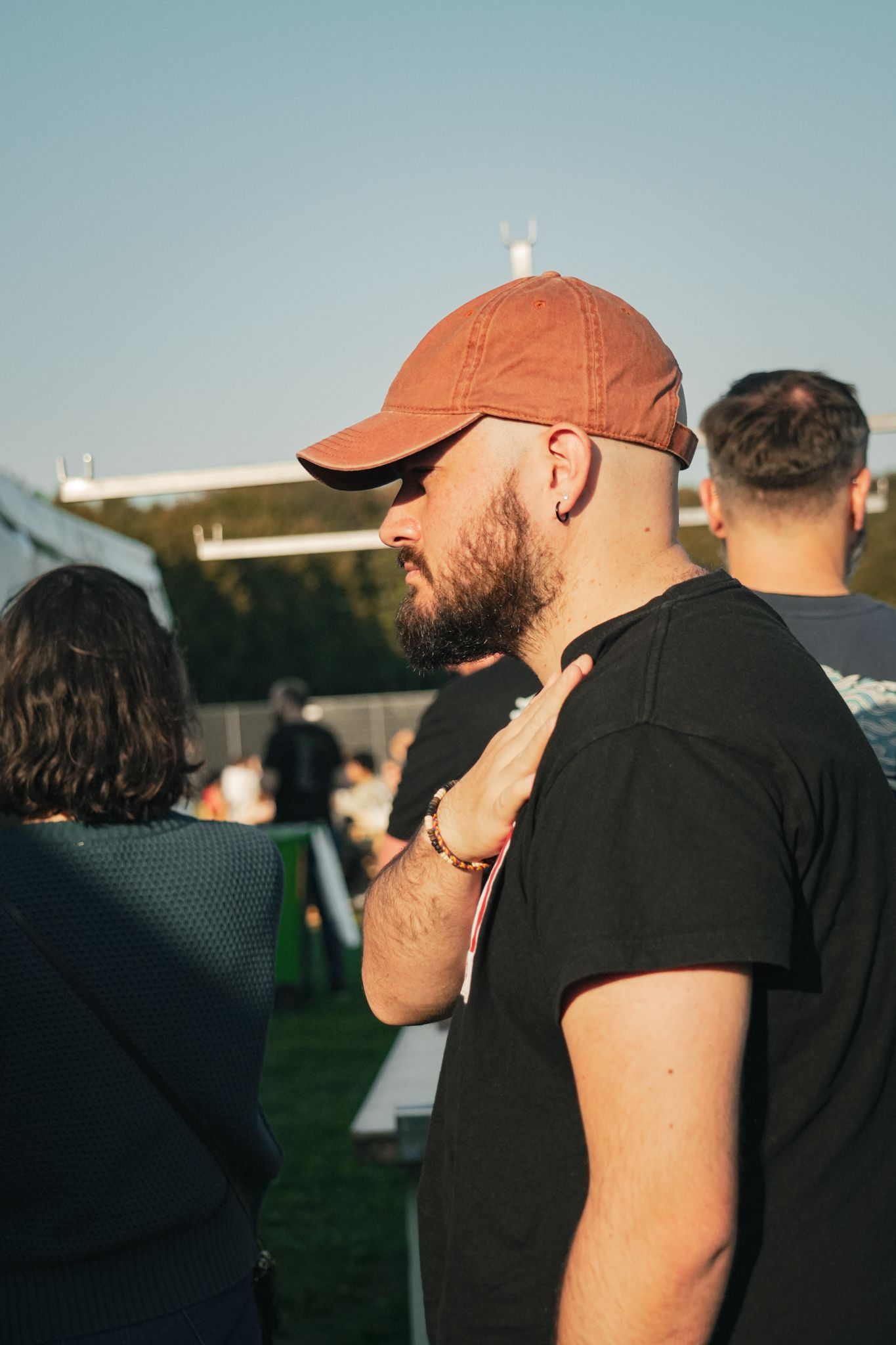 Man in orange cap and black shirt standing outdoors at a sunny event, seen in profile.