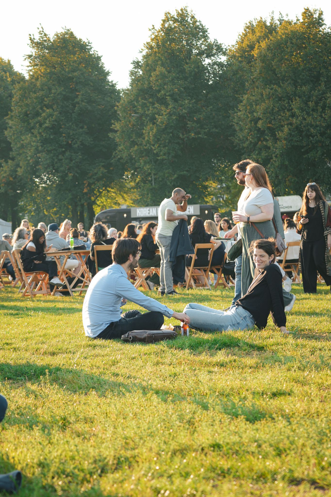 People sitting and standing on a sunny grassy lawn at an outdoor gathering with trees in the background