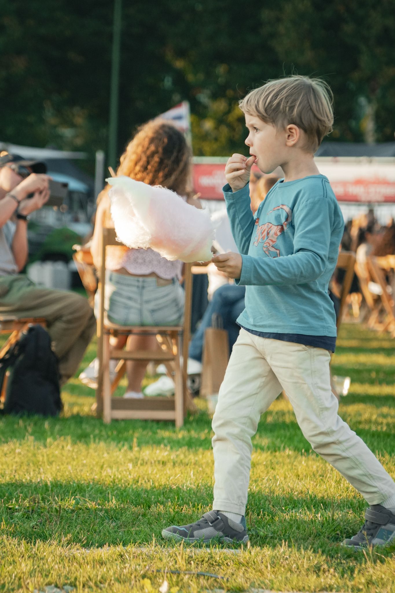Child walking on grass at an outdoor event, holding a drink with people seated in the background