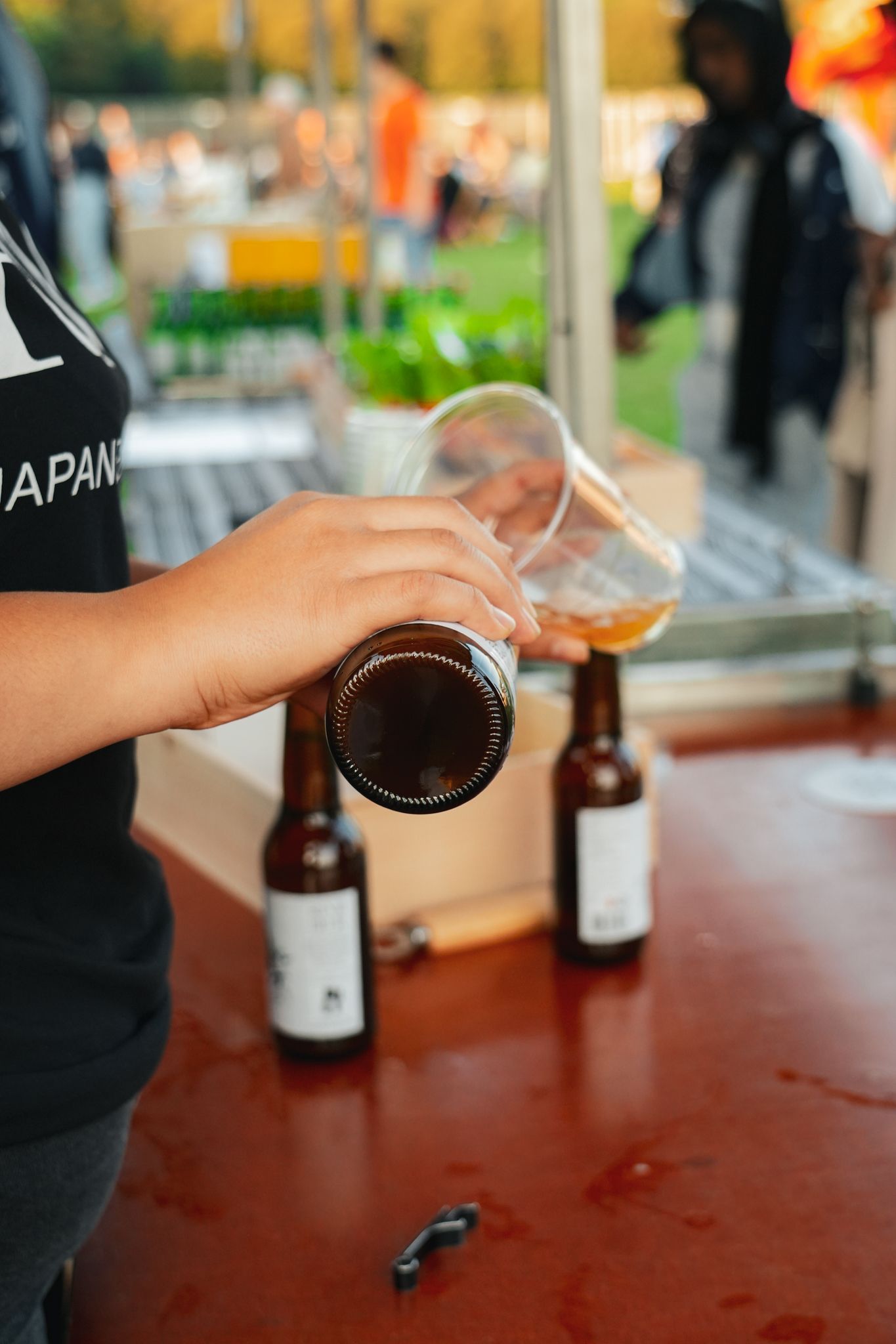 Hand pouring beer into a plastic cup beside two beer bottles on a red table outdoors