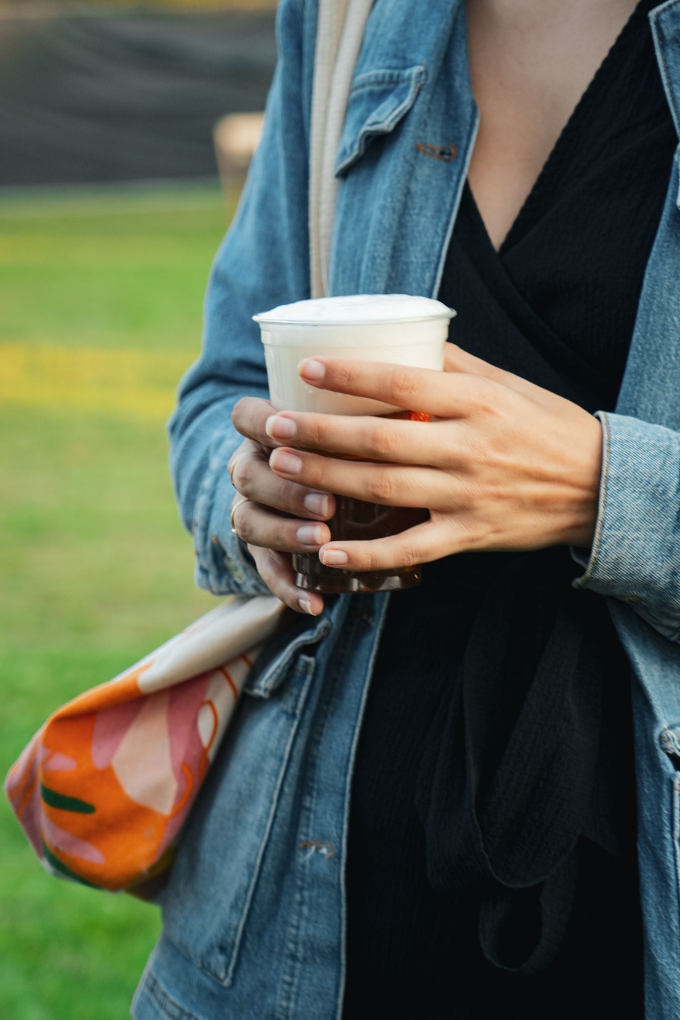 Person in a denim jacket holding a coffee cup outdoors, with a colorful bag at their side