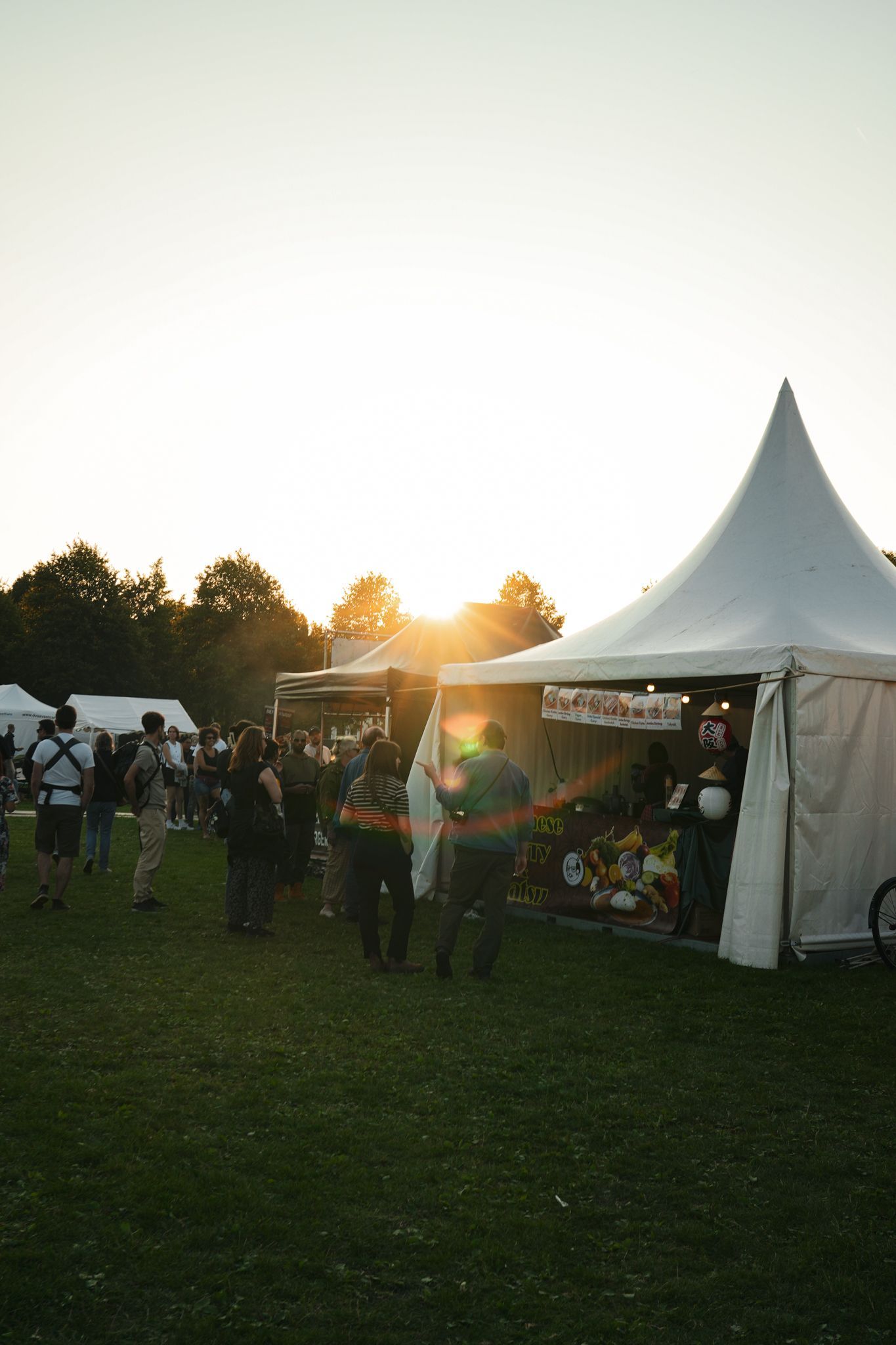 Outdoor festival at sunset with a white tent, people mingling, and the sun low over the grassy field.