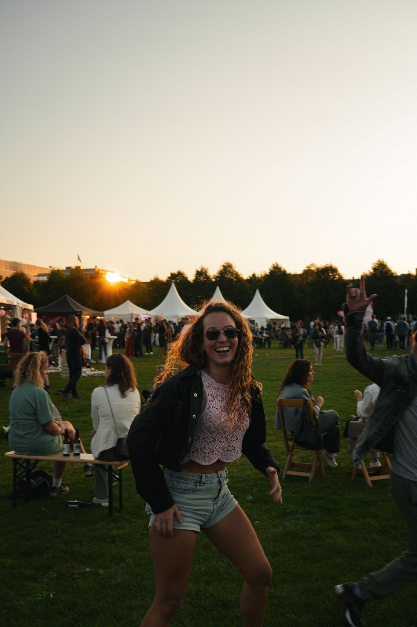 Woman dancing at an outdoor festival at sunset, with tents and a crowd in the background.