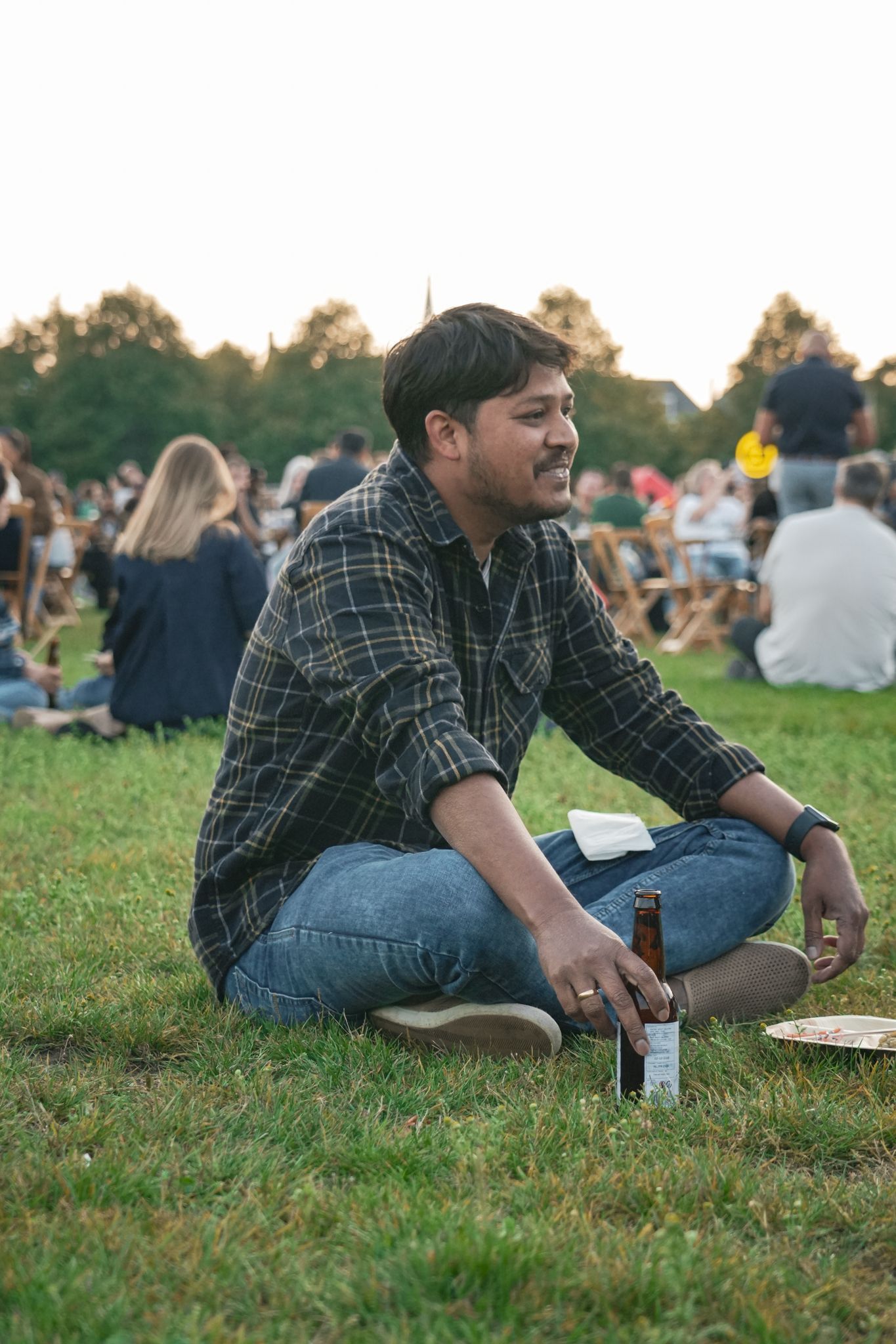 Man sitting cross-legged on grass at an outdoor gathering, holding a drink and smiling.