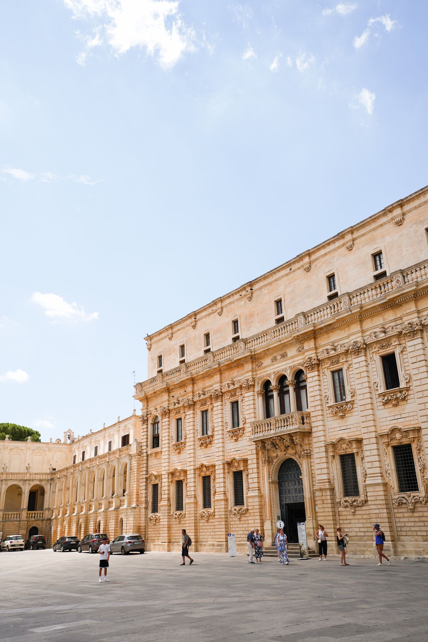 Historic stone courtyard with an ornate building under a blue sky, people walking nearby