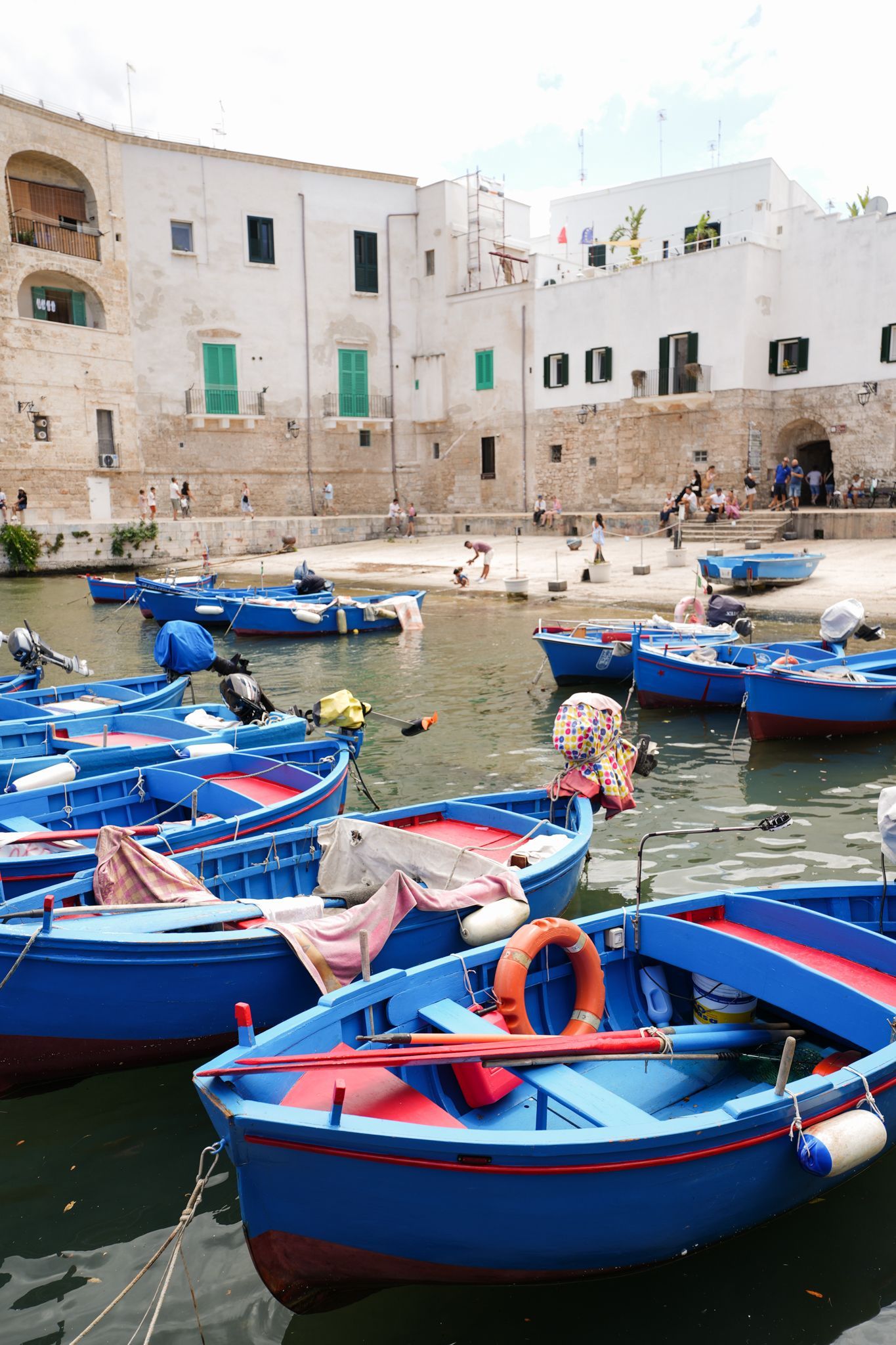 Blue boats floating in a small harbor beside white stone buildings and a sandy shore