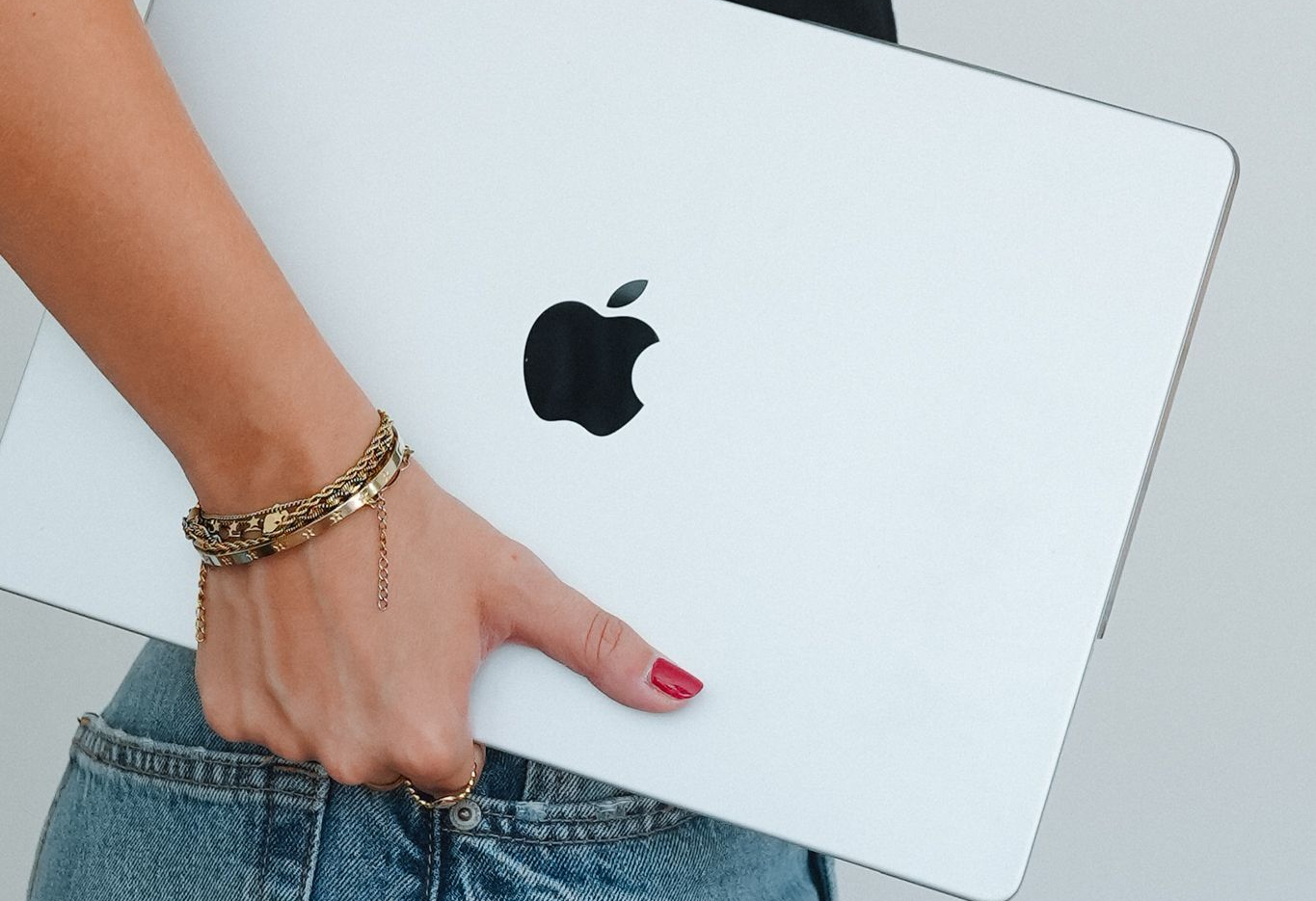 Hand holding a silver MacBook with a black Apple logo, wearing jeans and a gold bracelet