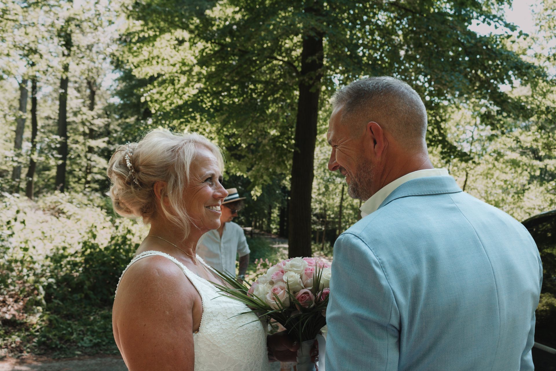 Two people face each other outdoors, one holding a bouquet of flowers in a wooded garden.
