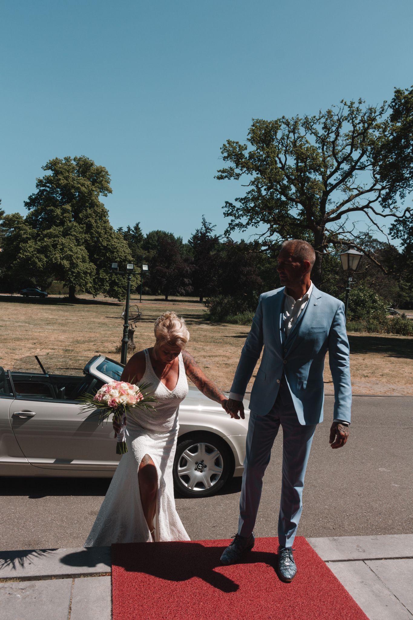Two people in formal attire walking on a red carpet outdoors beside a parked car.