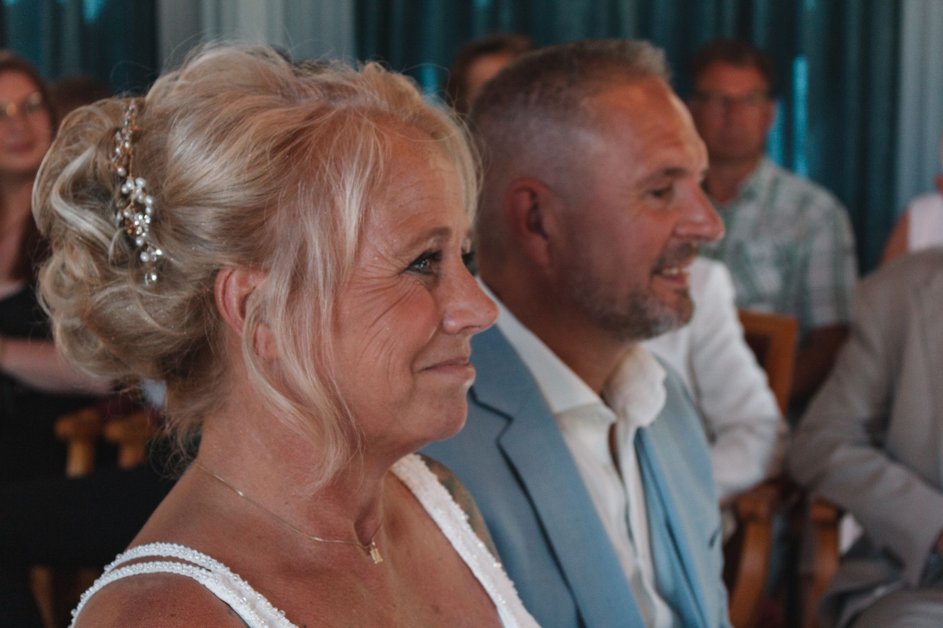 Two smiling attendees seated at an indoor event, with a woman in a white dress in the foreground