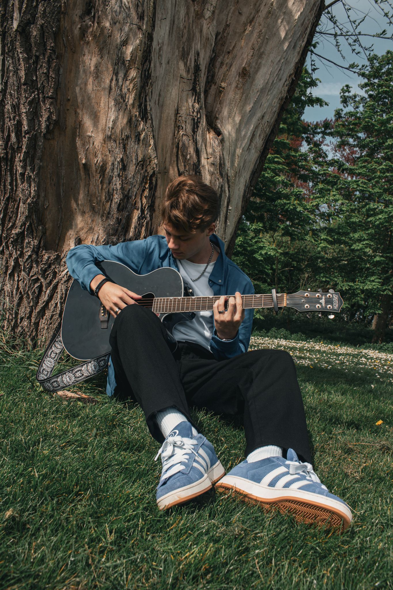 Person sitting under a large tree, playing acoustic guitar in a grassy park.