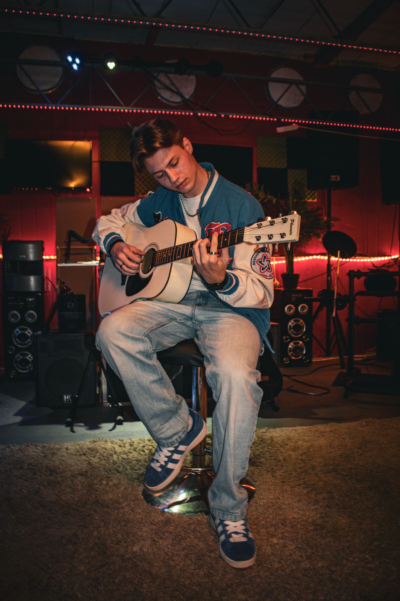 Person sitting and playing acoustic guitar in a dim red-lit studio setting