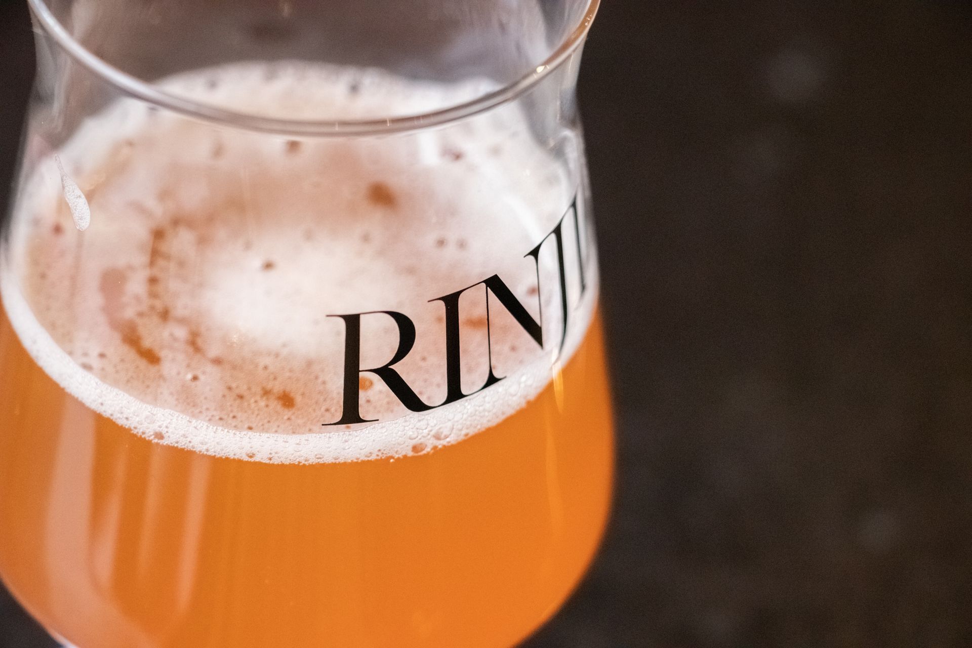 A glass of orange beer with a foamy head and partial black lettering on the rim.