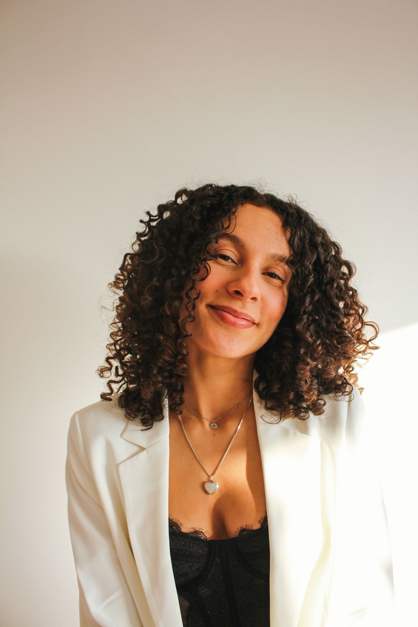 Woman with curly hair in a white blazer and black top, smiling against a plain wall.