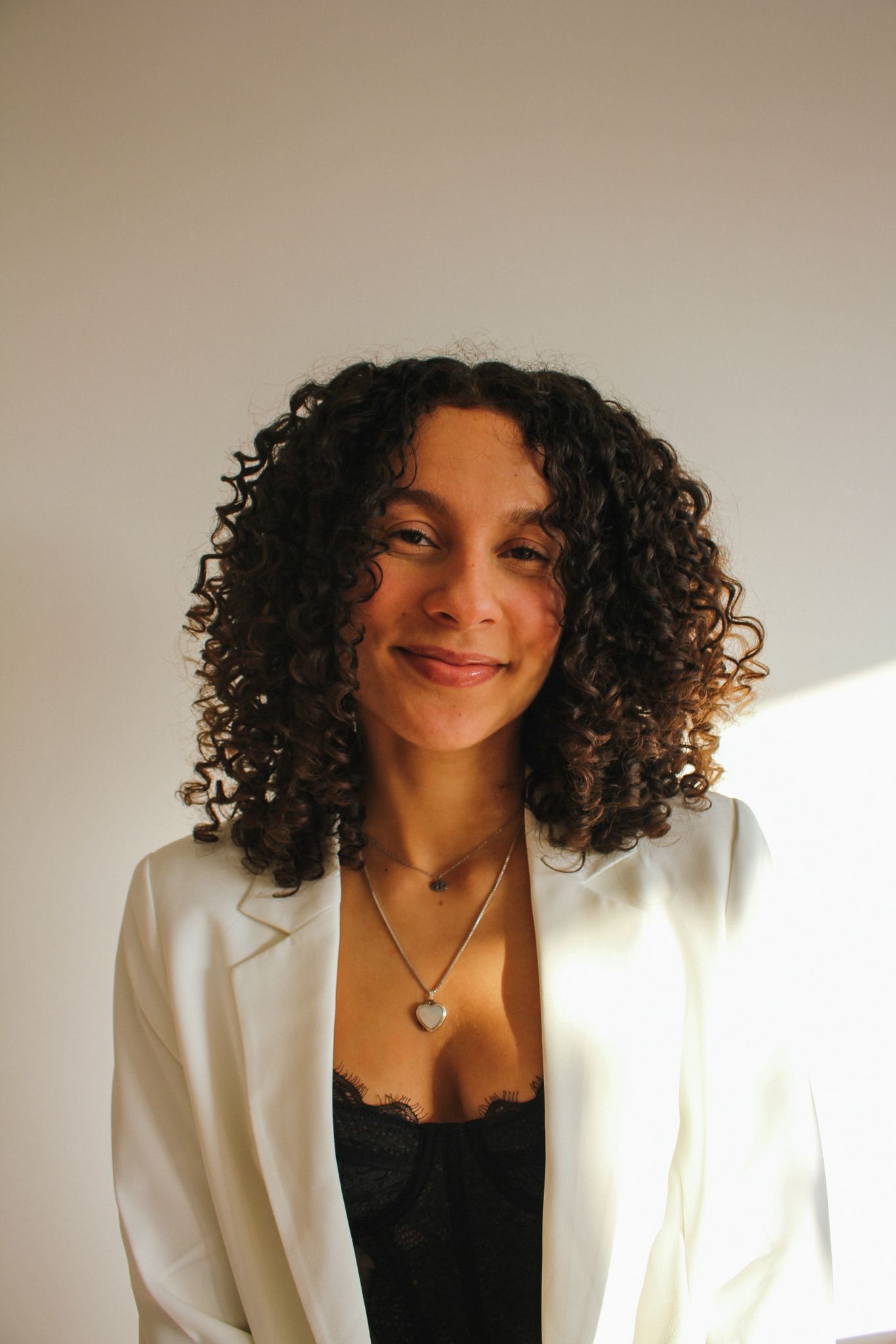 Woman smiling in a white blazer and black top against a plain wall with sunlight
